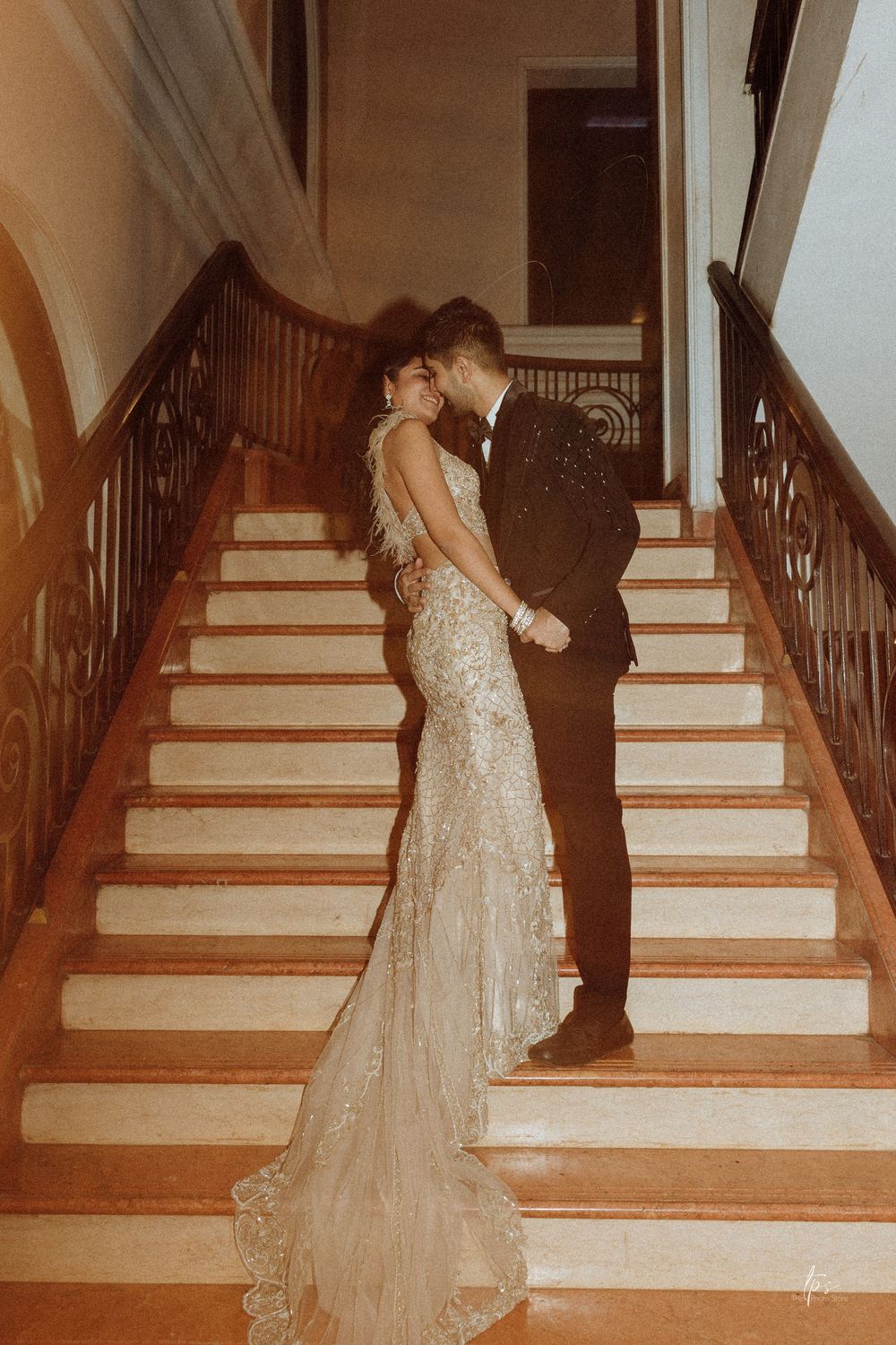 Bride in lace gown and groom in tuxedo share romantic moment on elegant staircase.