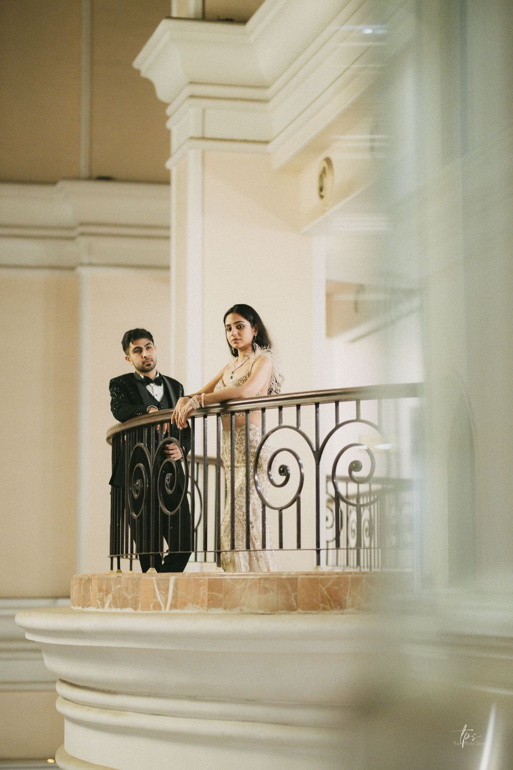 Elegant couple in formal attire standing on an ornate balcony with wrought iron railings in a grand venue.