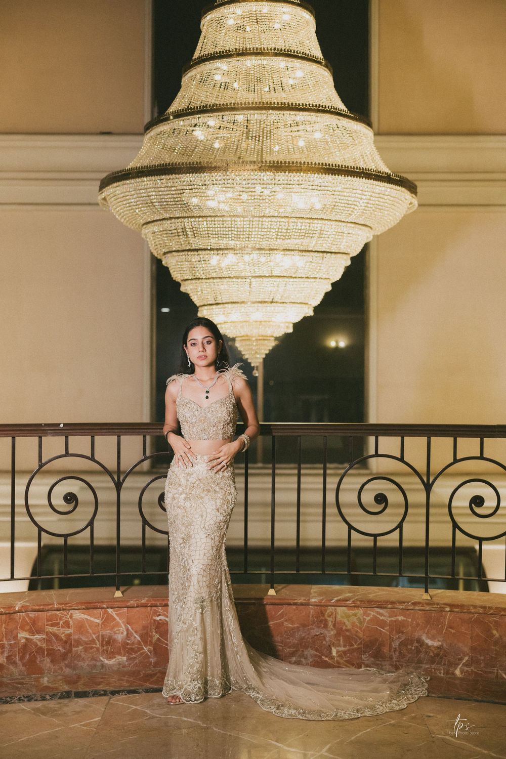Woman in gold embellished gown poses beneath grand crystal chandelier at elegant venue.