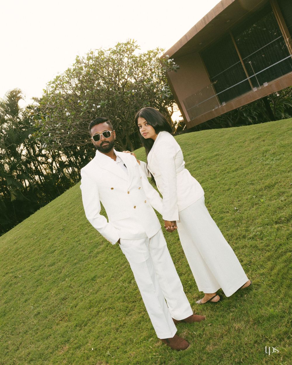 Couple in matching white suits holding hands on green lawn with modern building and trees in background.