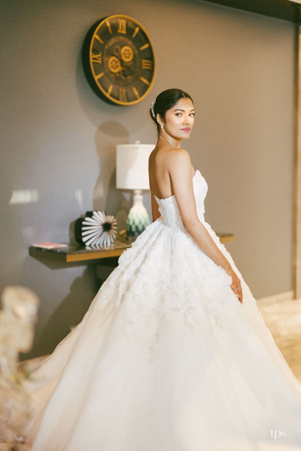 Elegant bride in a strapless white ball gown with lace details, posing gracefully in a softly lit room with a decorative clock.