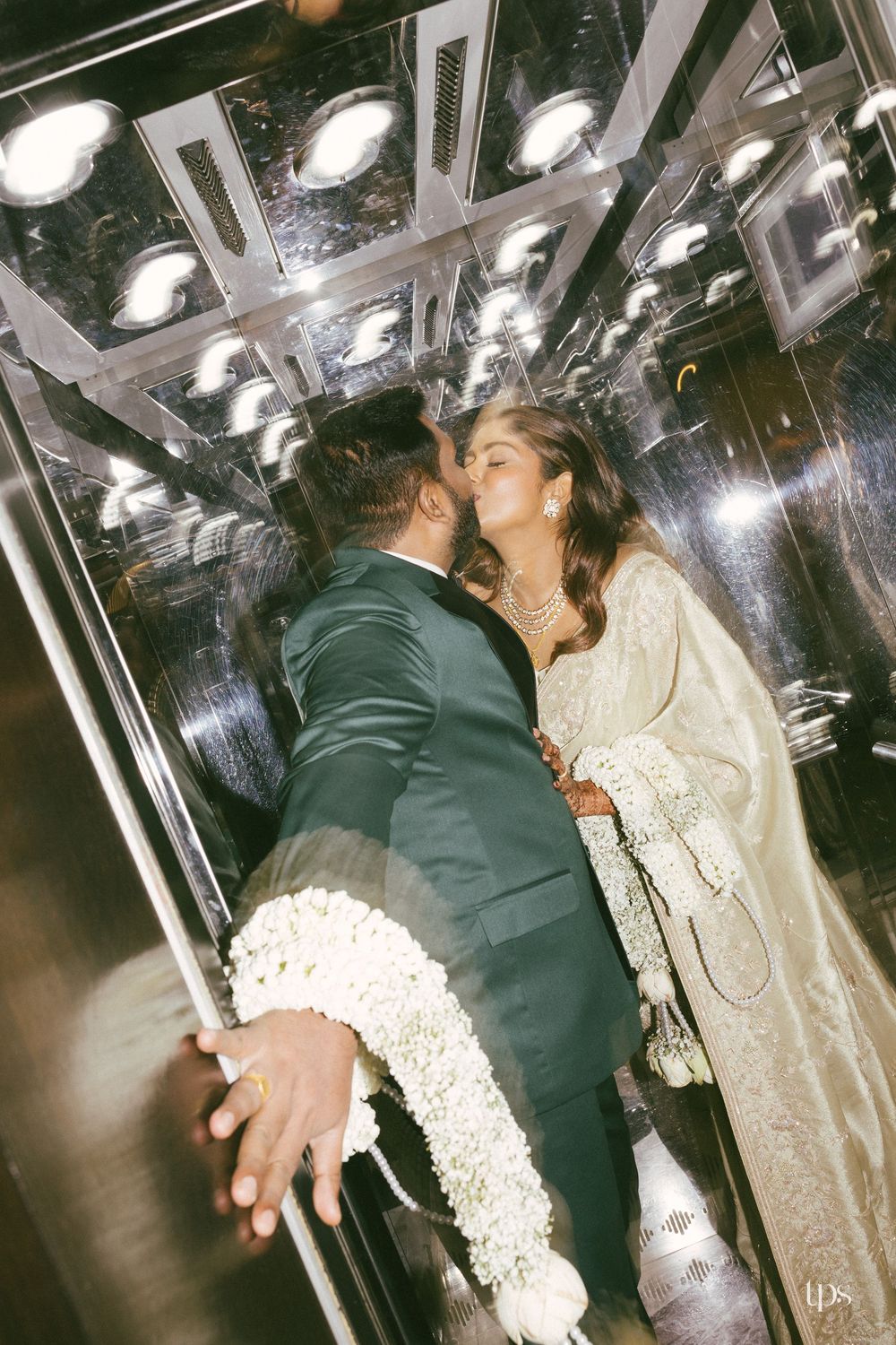 Couple sharing a romantic kiss in a mirrored elevator, bride in elegant white embroidered saree holding floral garland.