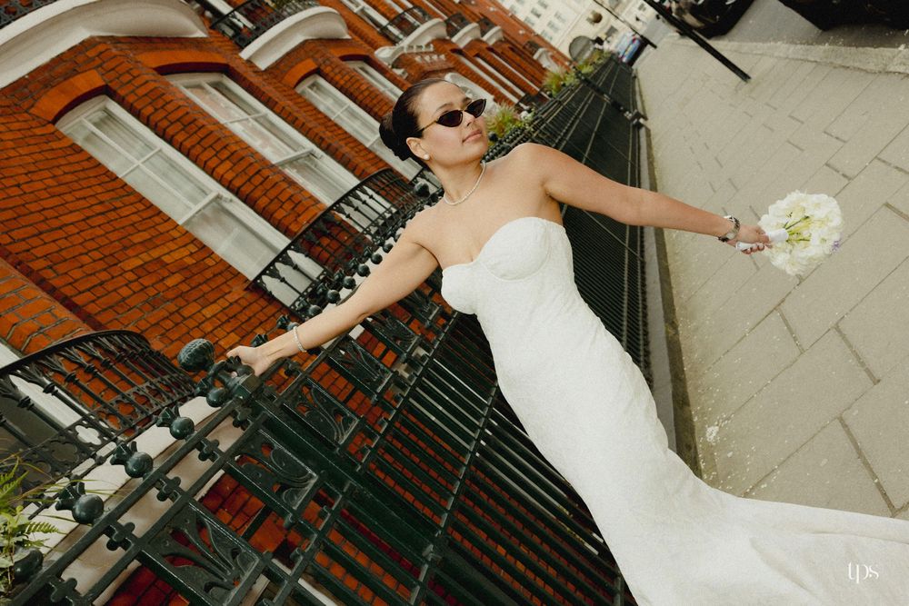 Bride in white strapless gown and sunglasses poses on balcony with bouquet, brick buildings in background.