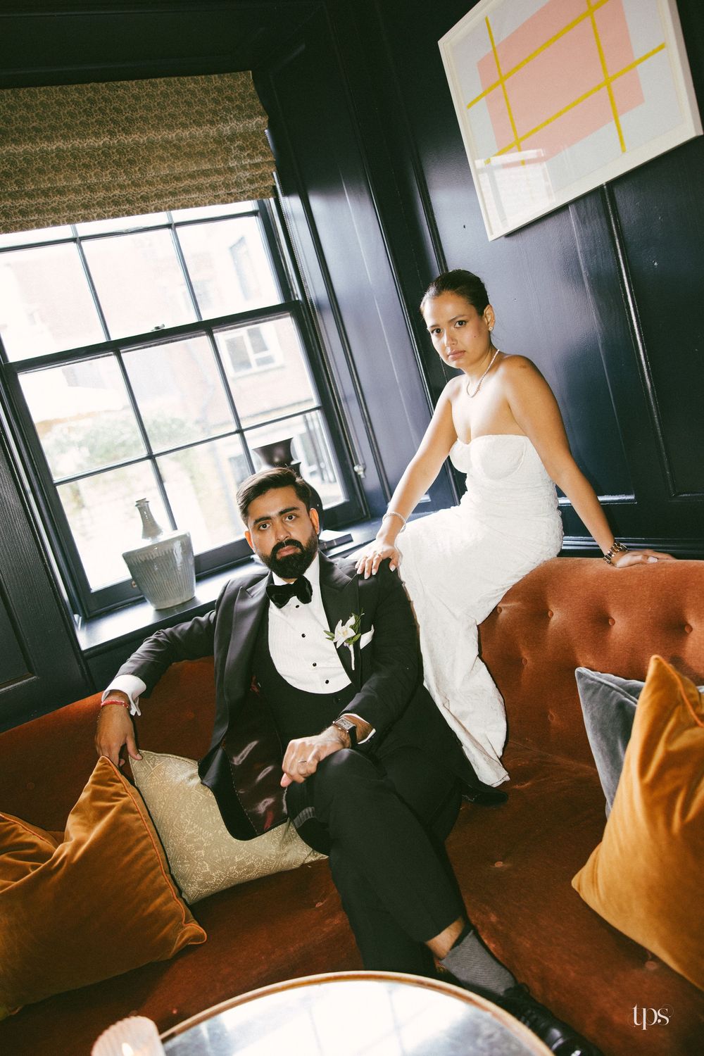 Elegant bride in white gown and groom in tuxedo posing on velvet sofa in a dark-walled luxury venue.