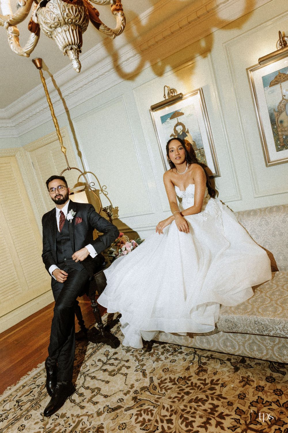 Bride in white ballgown sits on sofa while groom in black suit leans beside her in an elegant decorated room.