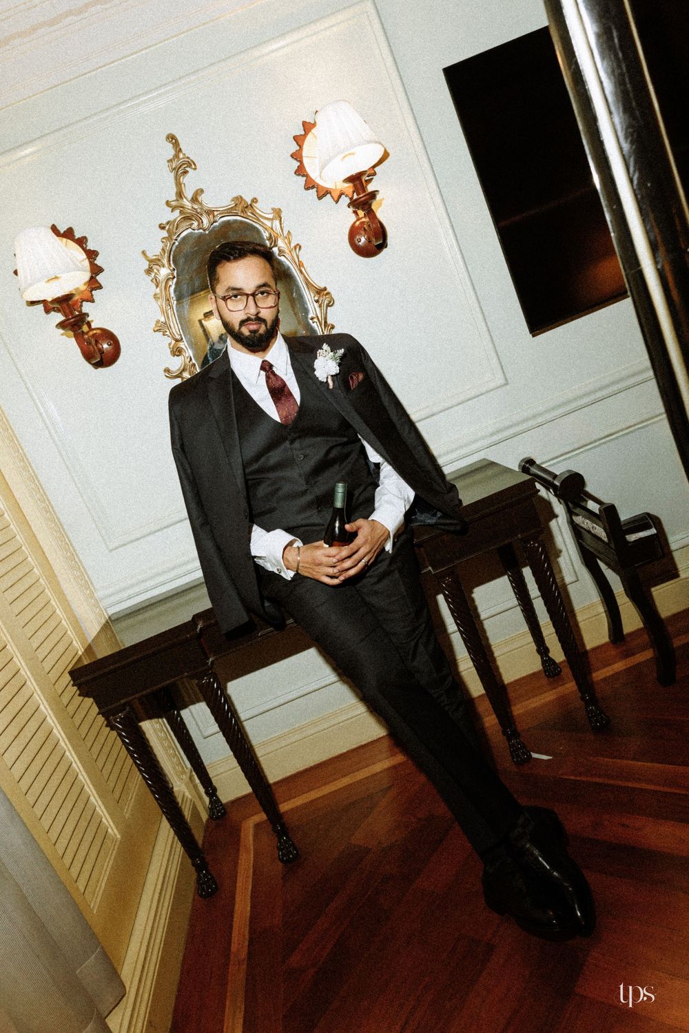 Dapper groom in black suit with burgundy tie holding wine bottle, posed elegantly in ornate venue interior.