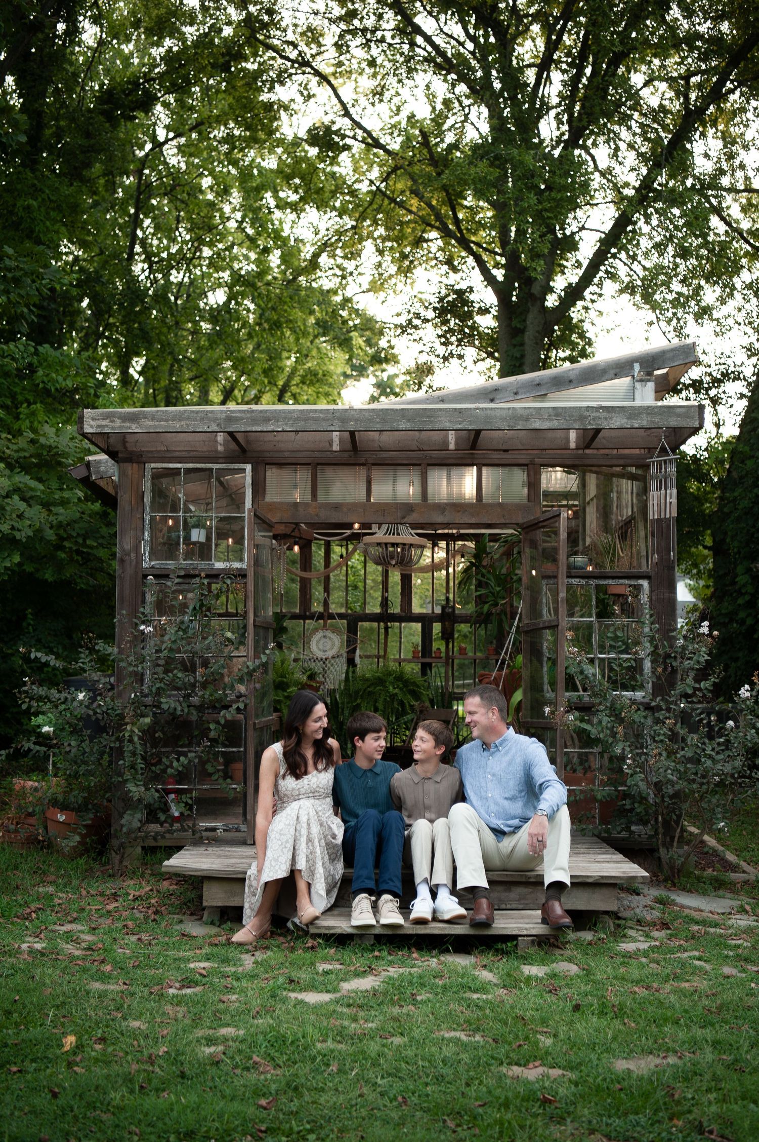 Family of four sitting together on steps of Nashville glass house  surrounded by lush green trees in a backyard garden.