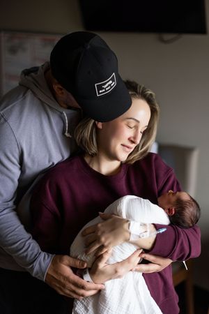 New parents tenderly hold their newborn baby, with mother in maroon sweatshirt and father in black cap kissing her head.