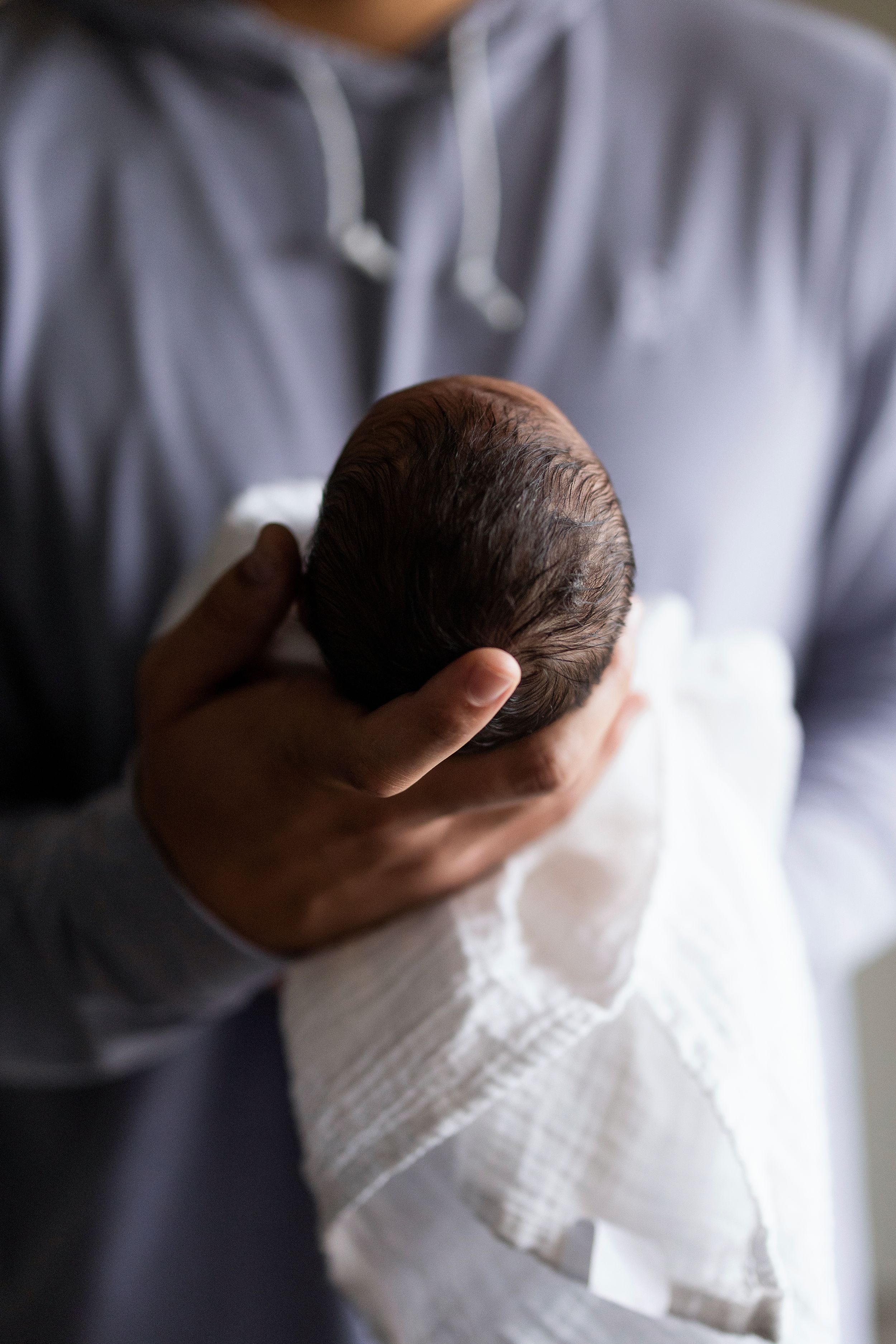 A parent in a gray hoodie gently craddles a newborn baby wrapped in a white muslin swaddle blanket.