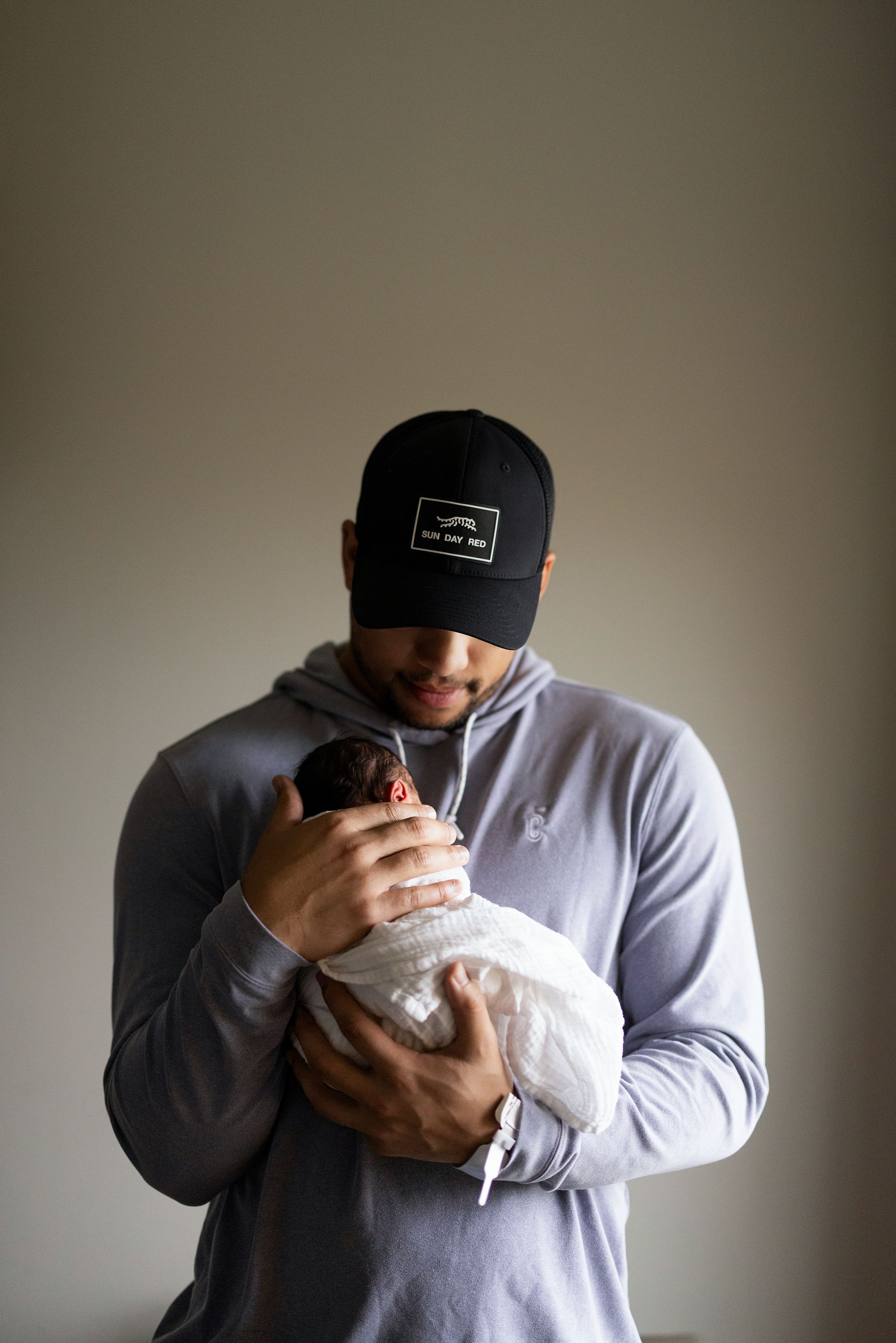 A man in a black cap and gray hoodie tenderly holds a swaddled newborn baby close to his chest.