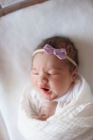 Newborn baby girl swaddled in white wrap, wearing purple bow headband, yawning with eyes closed.