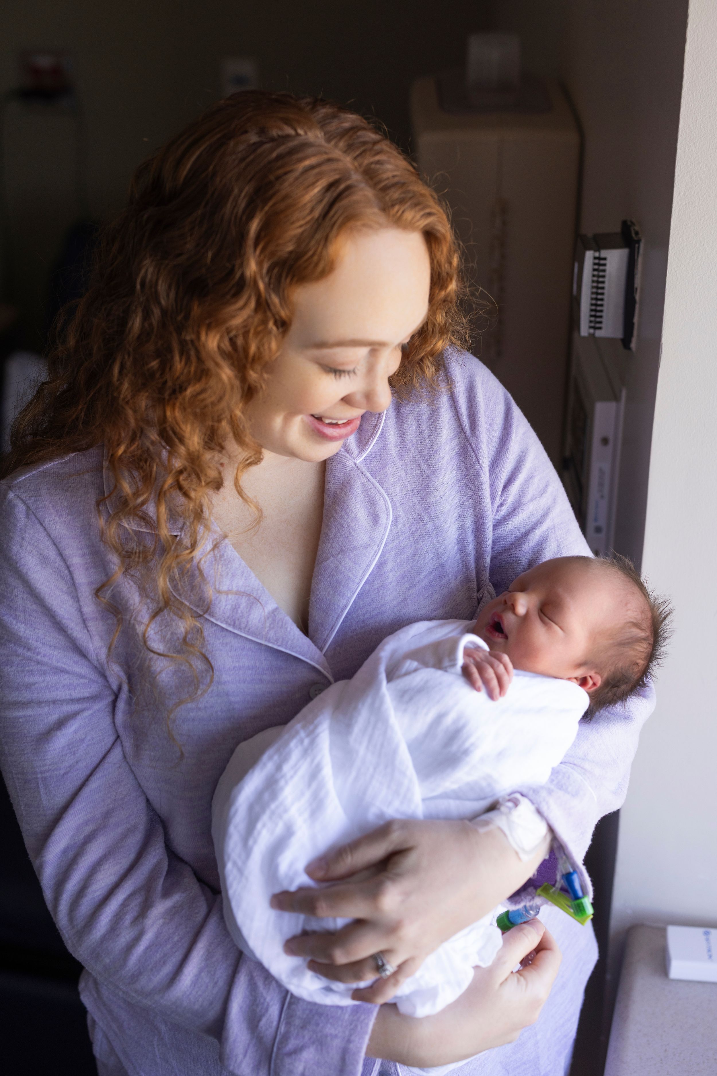 A smiling redhead woman in lavender pajamas cradles her swaddled newborn baby in a hospital room.