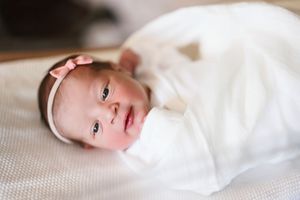 Newborn baby girl swaddled in white blanket wearing pink bow headband lying on soft surface.