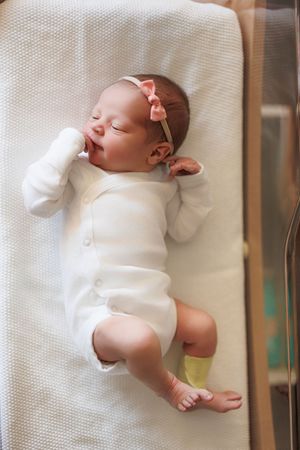 Newborn baby girl sleeping peacefully in hospital bassinet wearing white onesie and pink bow headband.
