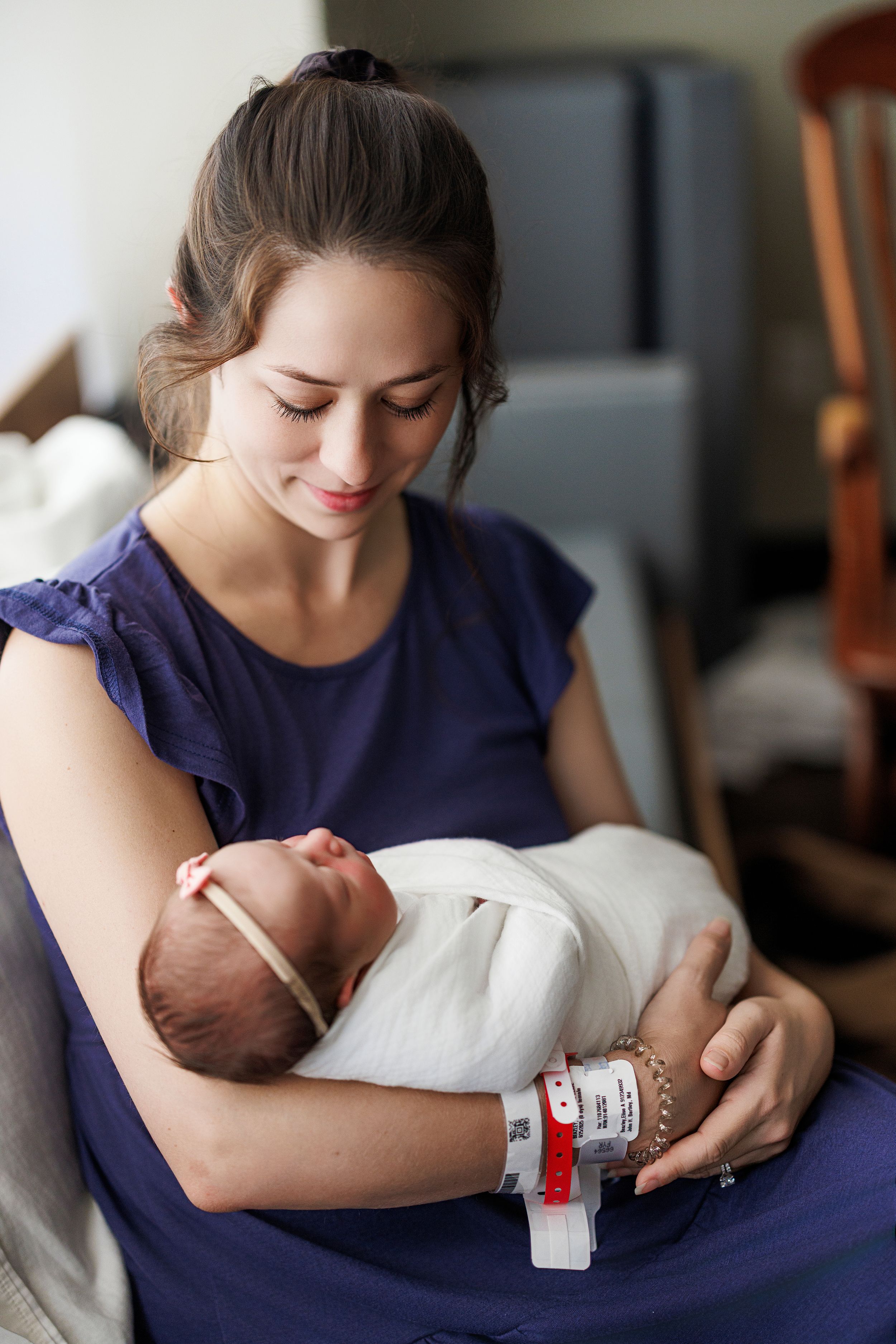 Young mother in blue dress tenderly holds swaddled newborn baby in hospital room after giving birth.