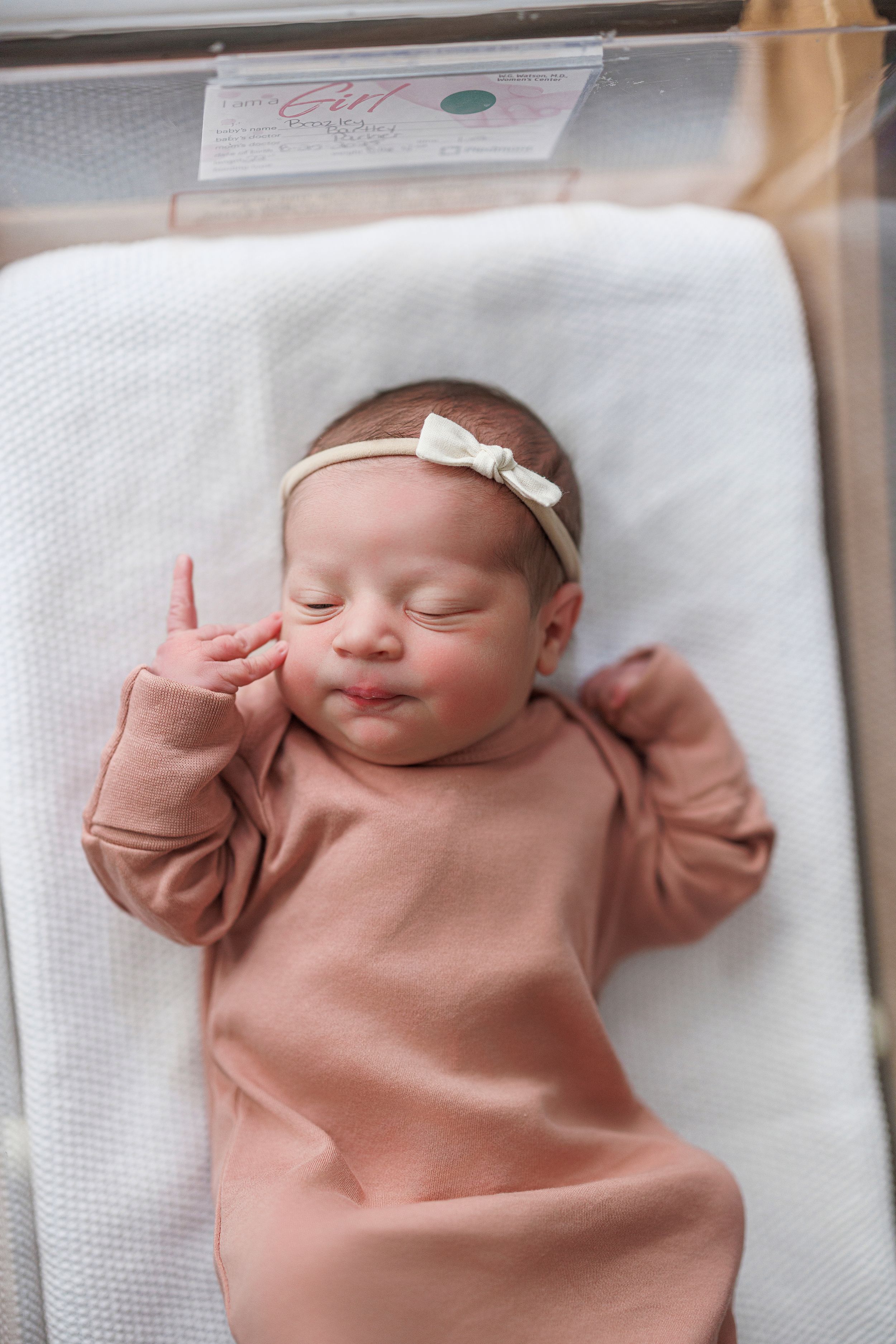 Newborn baby girl in pink gown and white bow headband sleeping peacefully in hospital bassinet.