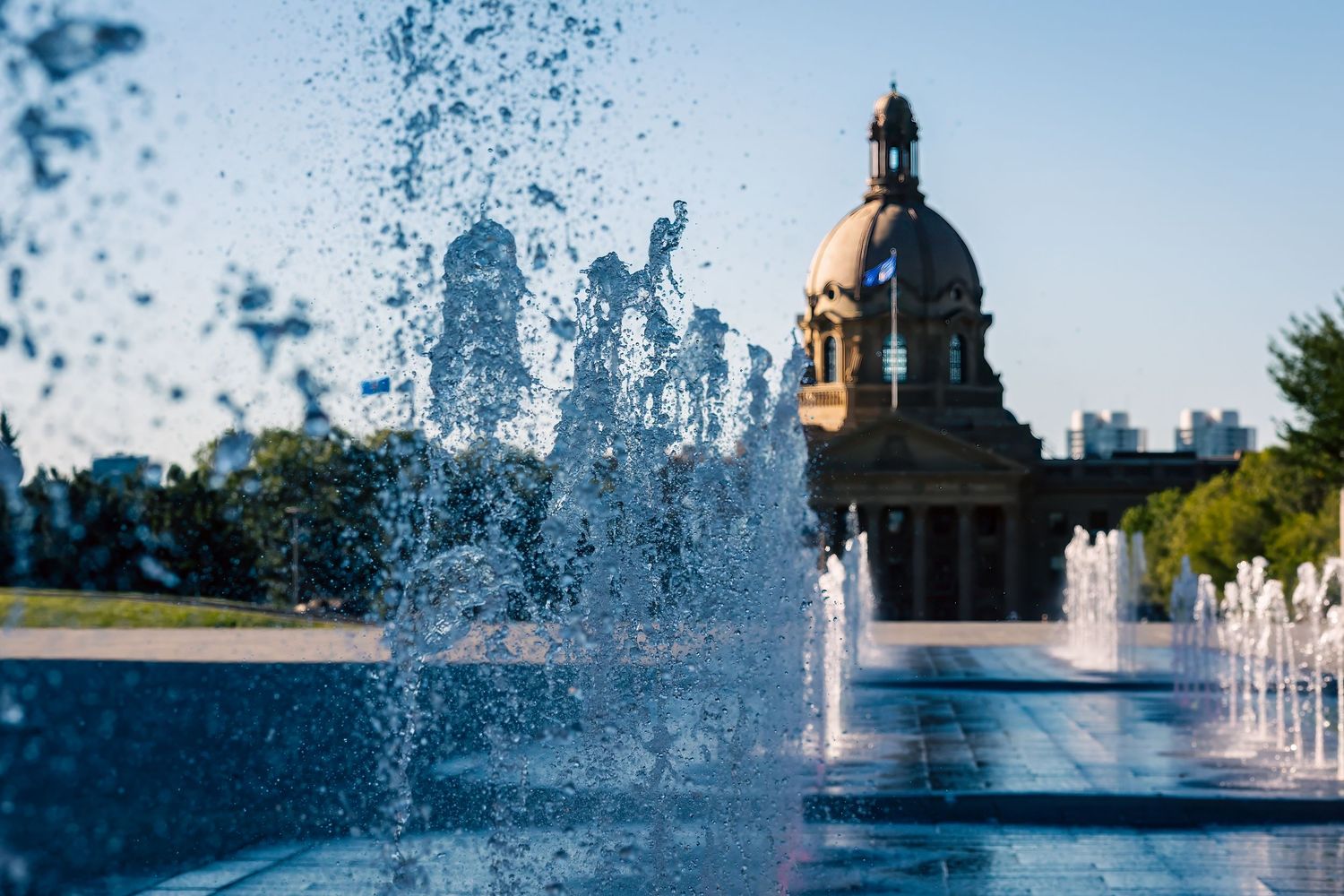 Water fountain splashing in front of the Alberta Legislature Building dome under a clear blue sky.