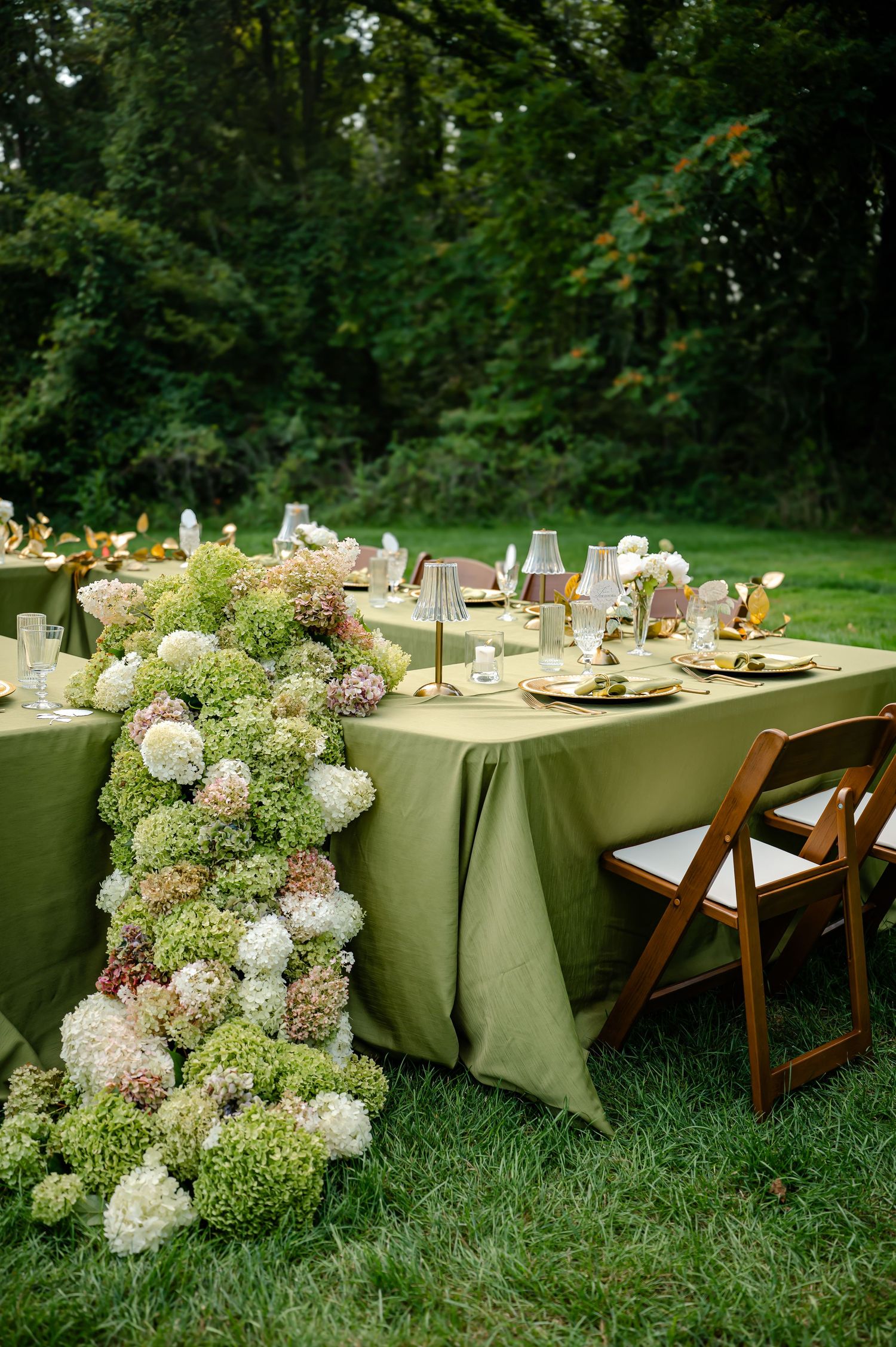 Elegant outdoor wedding reception table with green linen, hydrangea floral runner, gold accents, and wooden folding chairs on grass.