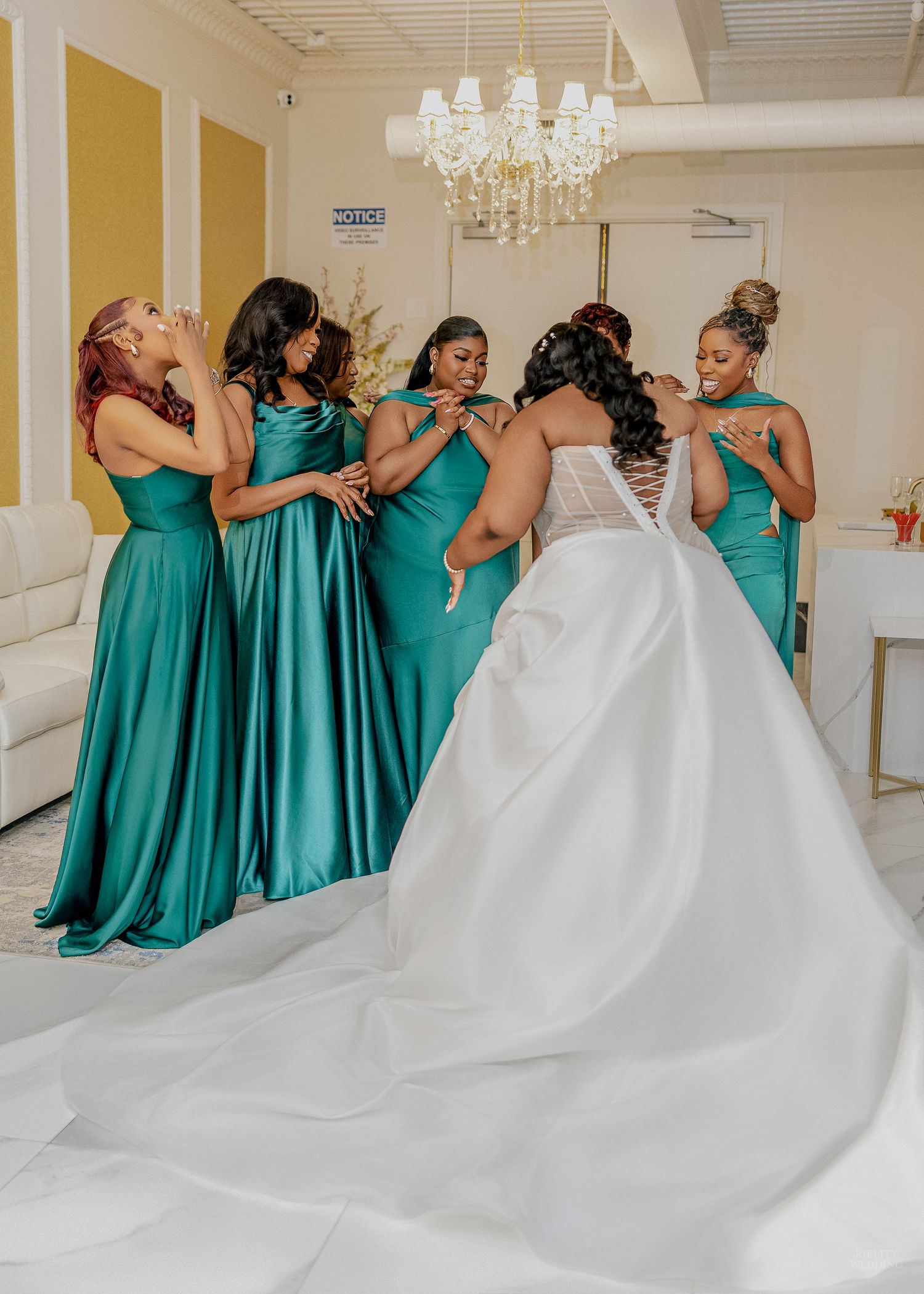 Bride in white gown with bridesmaids in teal dresses preparing together in elegant room with chandelier.