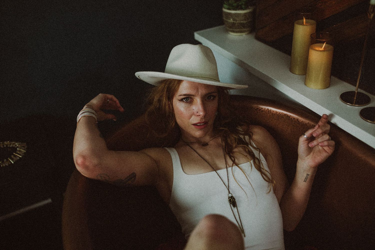 Woman in white bodysuit and wide-brim hat reclines on leather chair near candles in a dark moody setting.