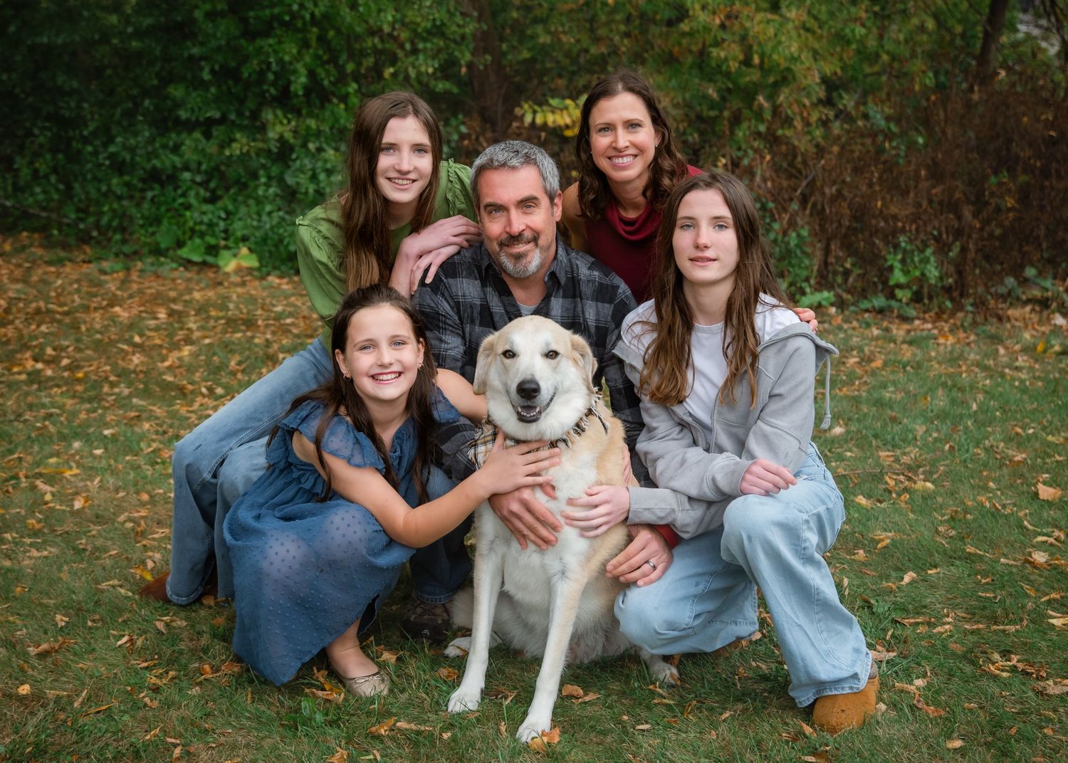 Family of five smiling outdoors in autumn with a happy yellow Labrador dog, surrounded by fall foliage.