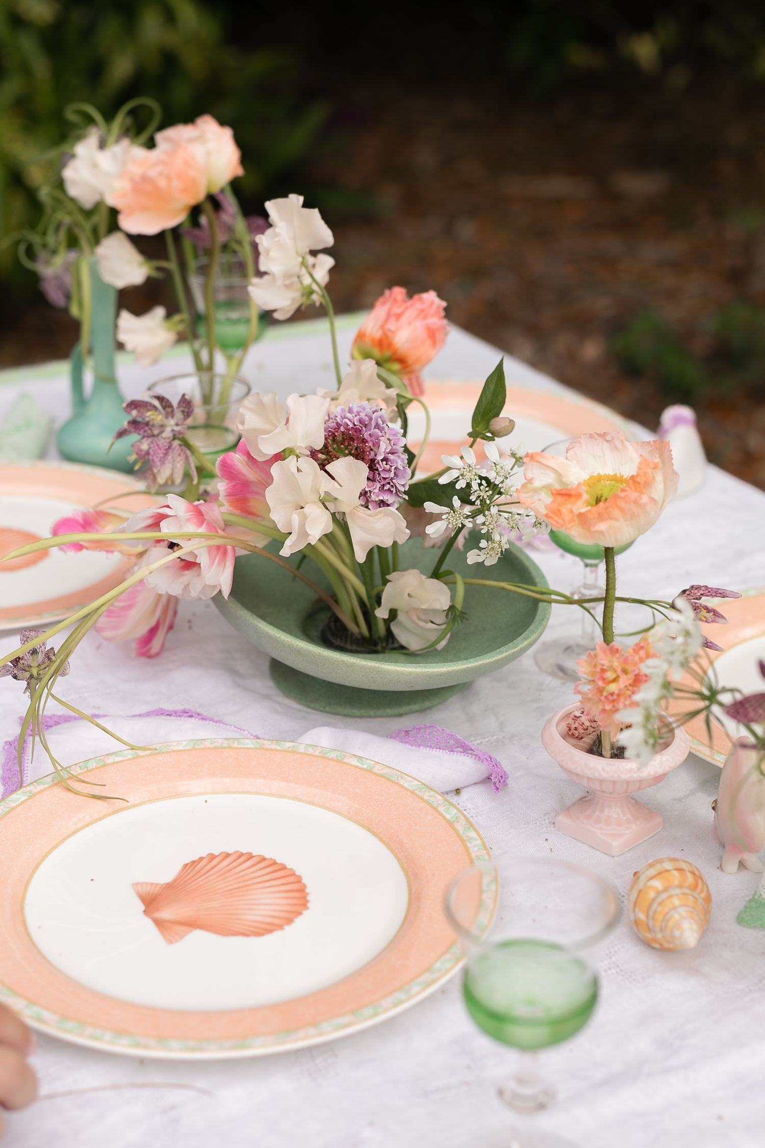 Elegant outdoor table setting with floral centerpiece, seashell plates, pastel colors, and green glassware.
