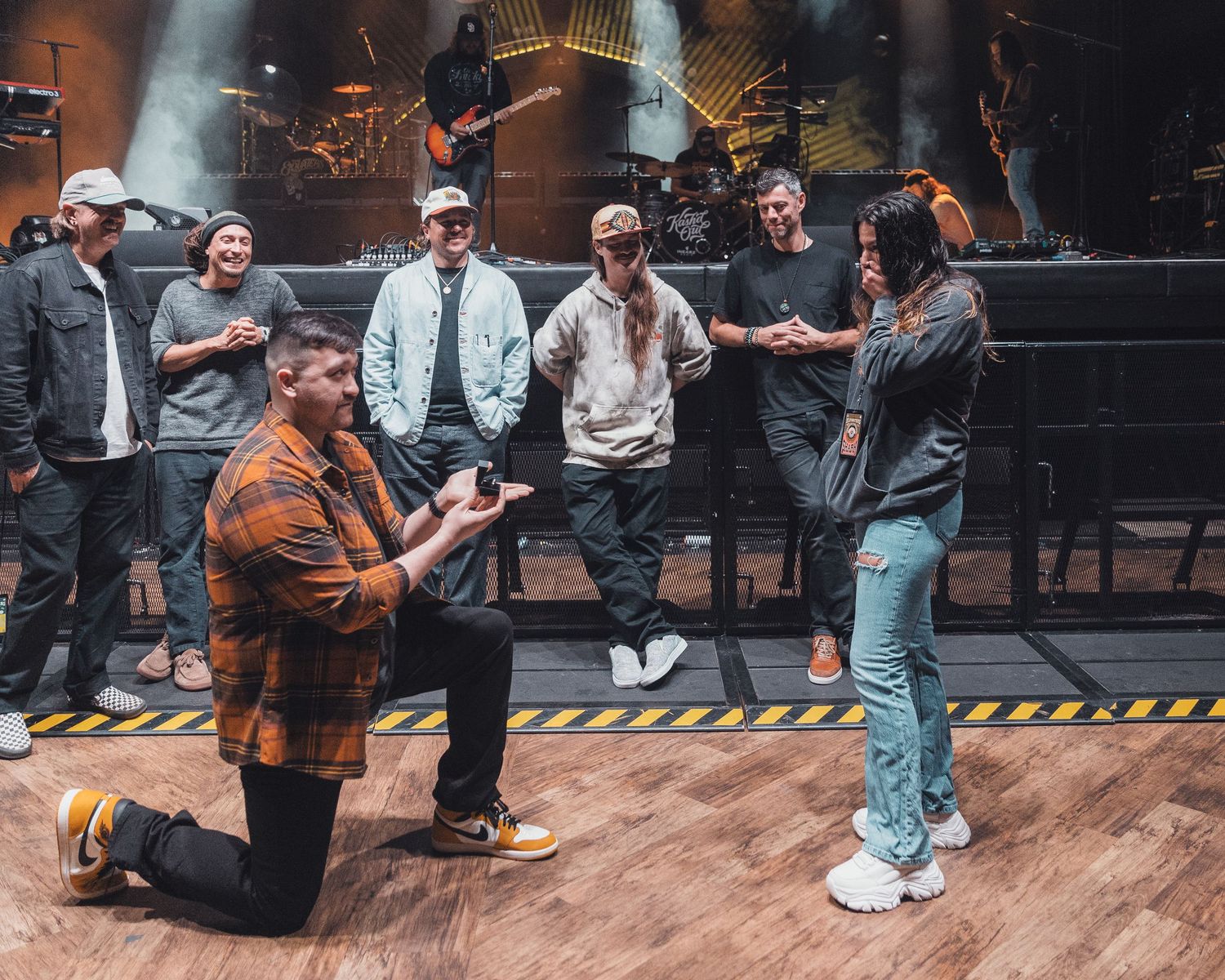 Man proposes on one knee to woman on stage while group of friends watches during a concert event.