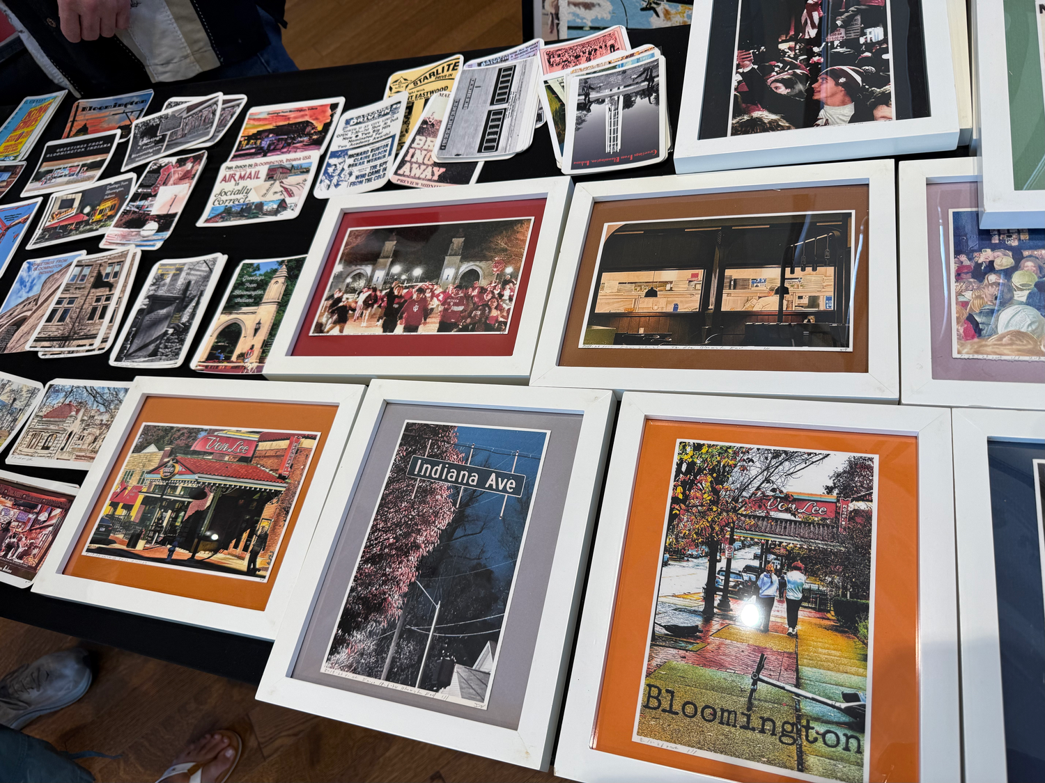 A table display of matted art prints and postcards featuring colorful city scenes including Bloomington at an art fair.