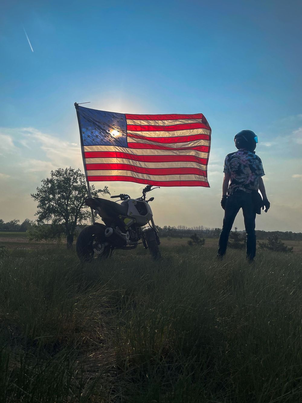 Person standing beside a motorcycle with an American flag waving at sunset in an open field.