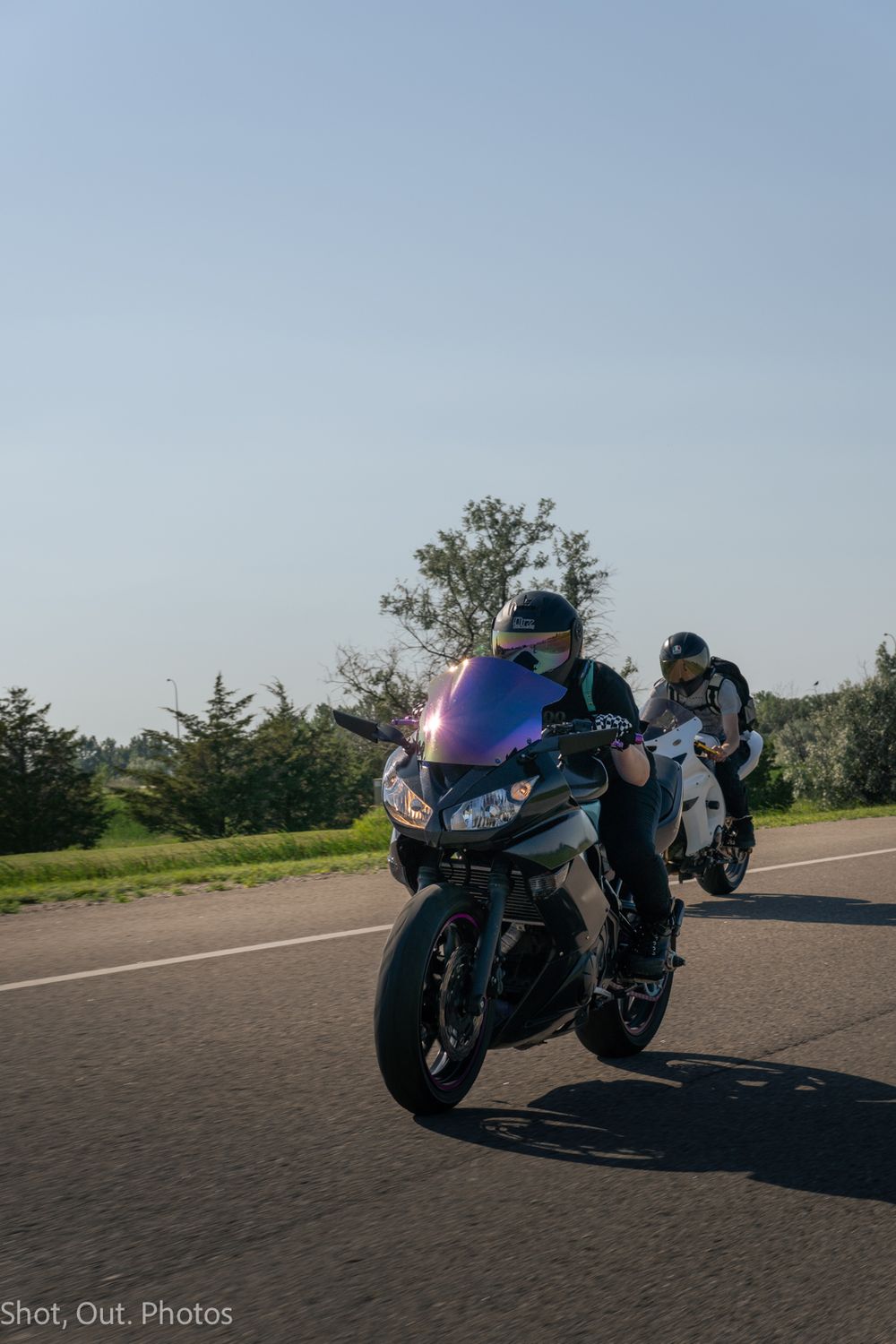 Two motorcyclists riding sport bikes on an open road surrounded by green trees under a clear blue sky.