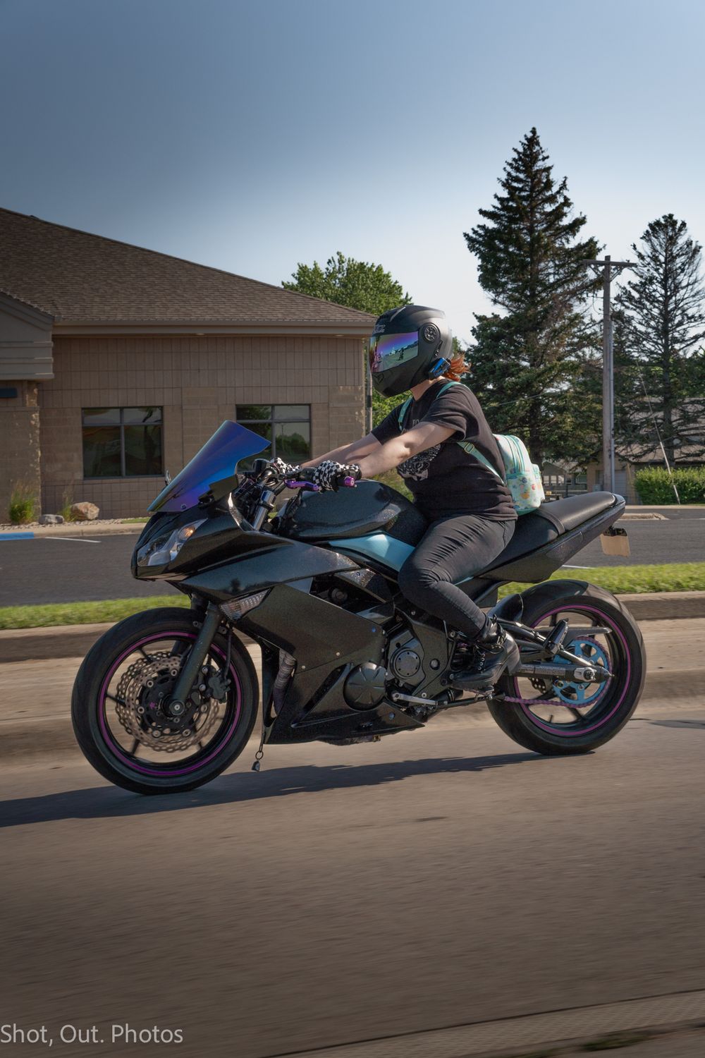 A motorcyclist in a helmet rides a dark sport bike on a road past a building and pine tree.