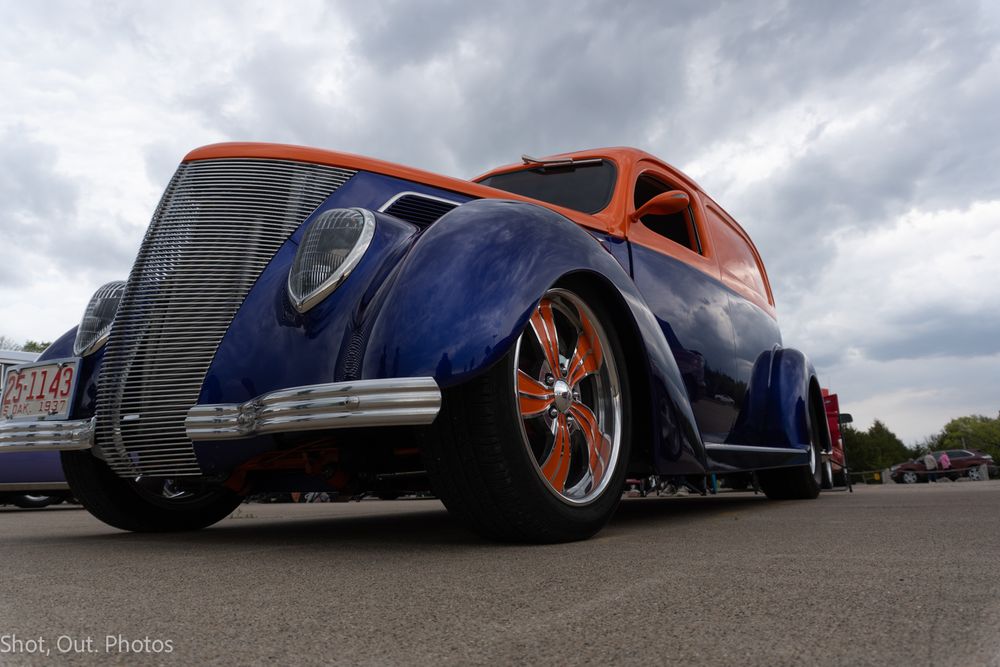 Custom blue and orange classic hot rod car with chrome wheels at an outdoor car show under cloudy skies.