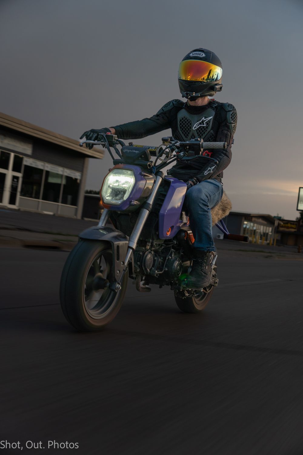 A motorcyclist in a reflective helmet and riding gear performs a stunt on a small motorcycle at dusk in a parking lot.