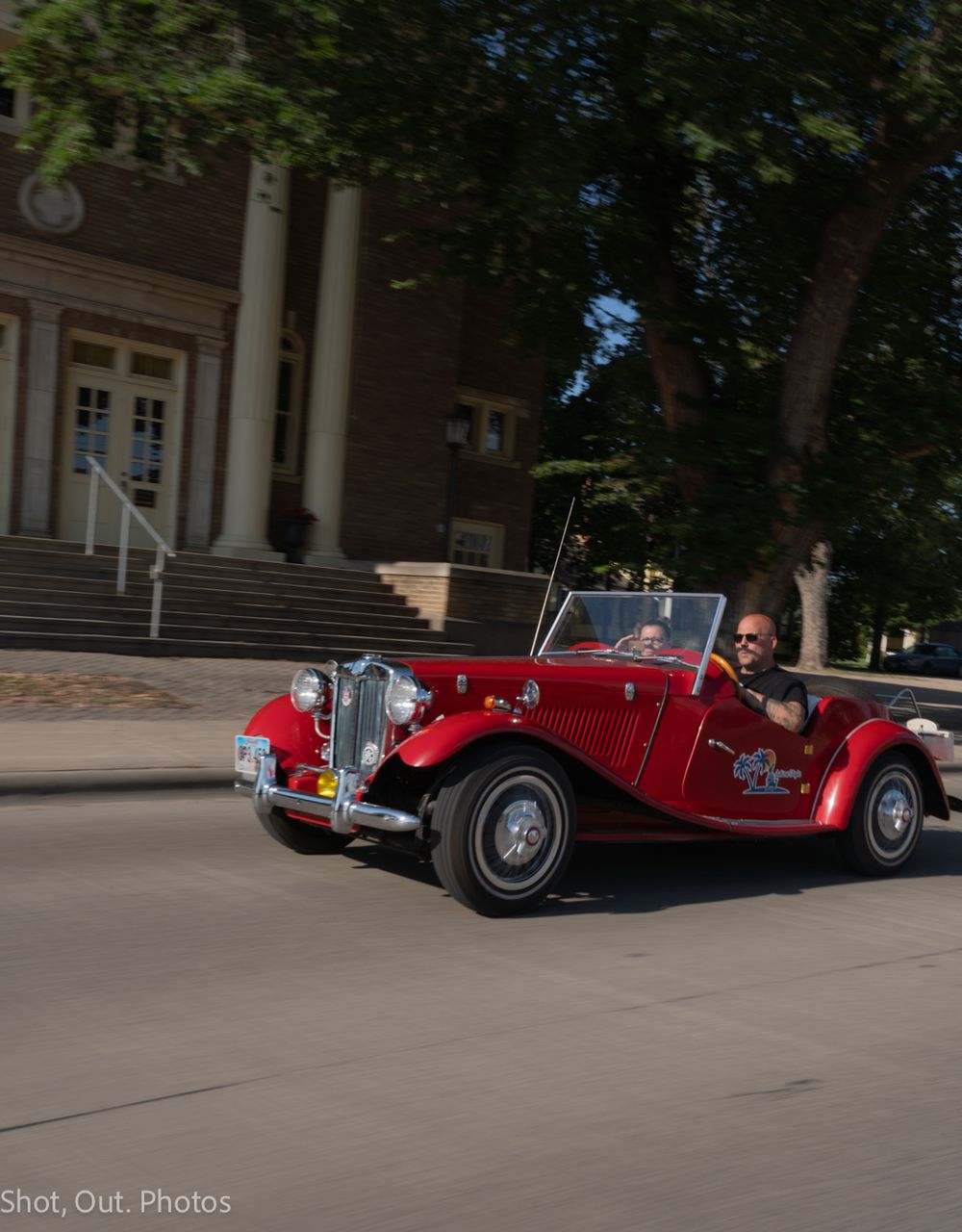 A man drives a classic red MG TD convertible sports car past a historic building on a sunny day.