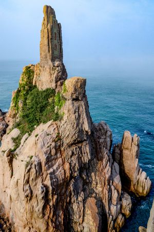Dramatic rocky sea stack formation rising from coastal cliffs with lush greenery, overlooking blue ocean waters on a misty day.