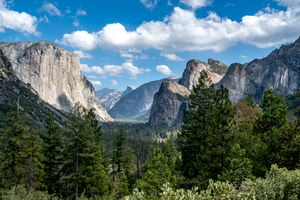Granite cliffs and rocky peaks rise above a lush evergreen forest in a scenic mountain valley under a bright blue sky with white clouds.