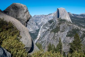 Half Dome rises above a granite valley in Yosemite National Park, seen from Glacier Point past a large boulder and evergreen trees.
