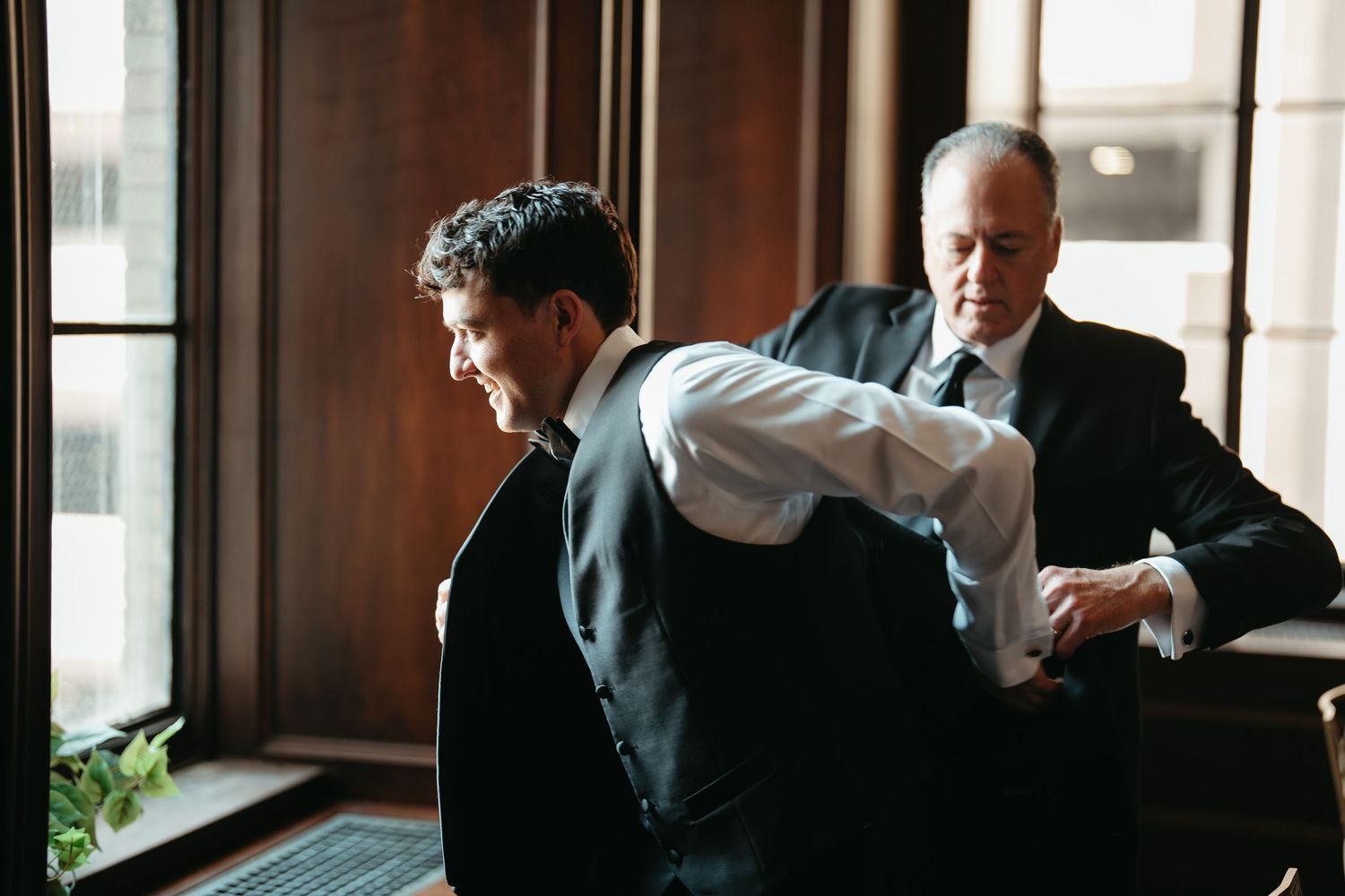 Groom being helped into his vest by groomsman near window before wedding ceremony.