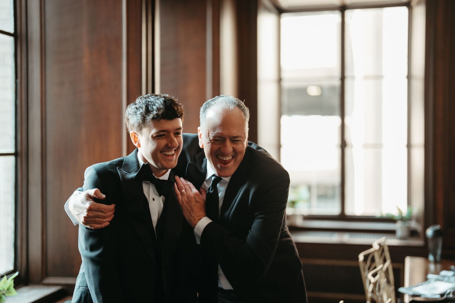 Two men in tuxedos share a joyful embrace, laughing together in an elegant wood-paneled room.