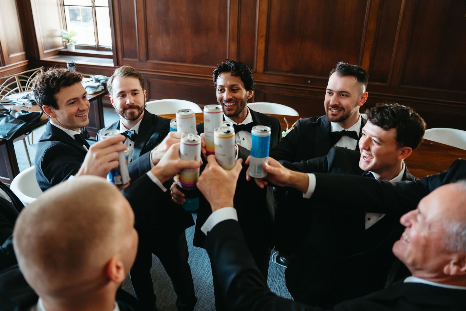 Groomsmen in black tuxedos toasting with canned drinks at a wedding celebration indoors.