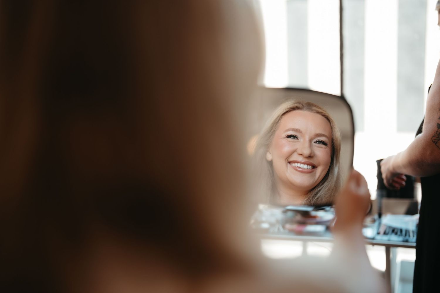 Smiling blonde woman getting makeup done, reflected in a vanity mirror with bright lights in a salon.