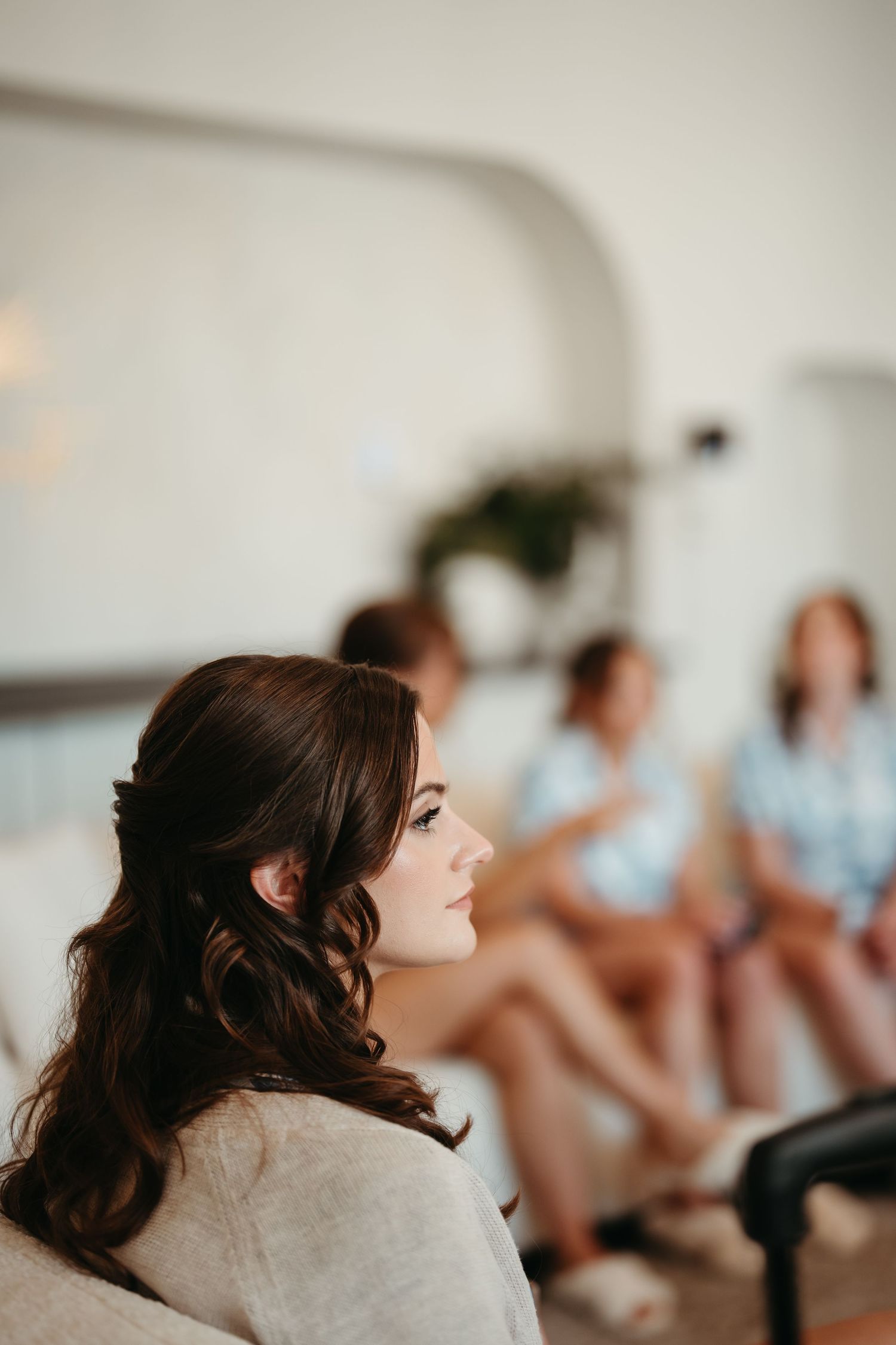 Bride with elegant half-up curled hairstyle sits in foreground while bridesmaids in blue robes chat in blurred background.