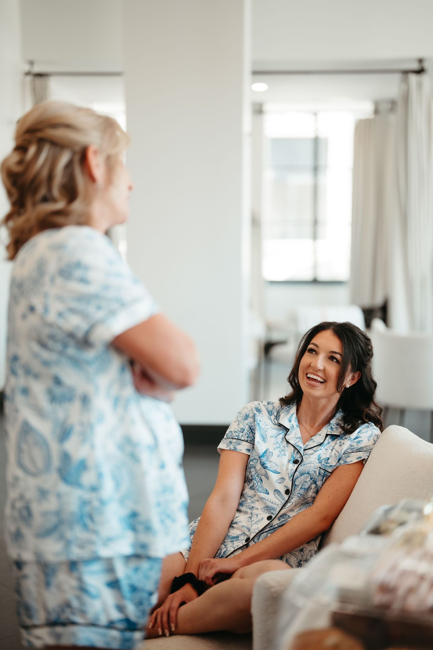 Two women in matching blue floral pajamas laugh together in a bright white room during a bridal getting ready session.