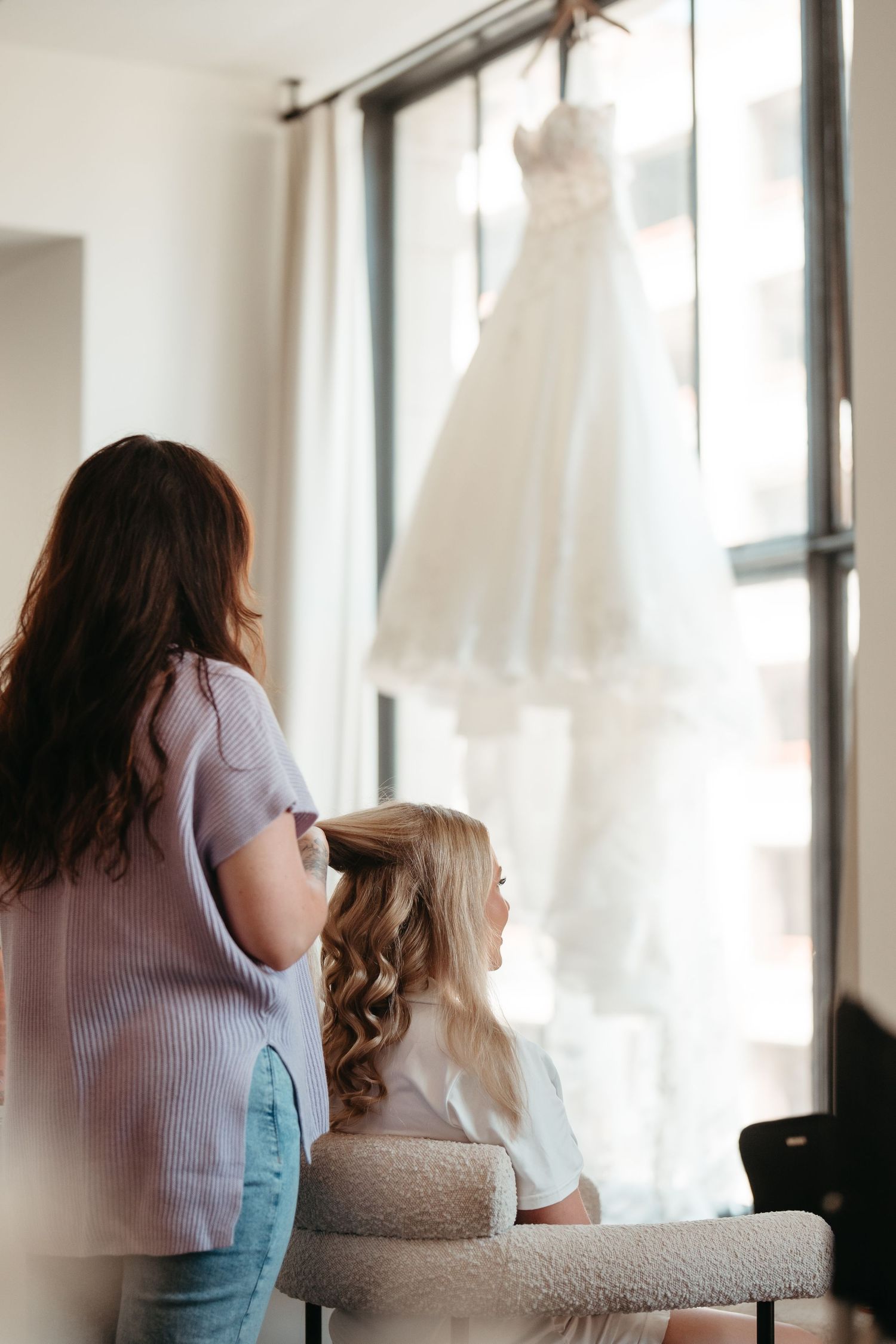 Woman and young girl admire a white wedding dress hanging by a bright window before the ceremony.