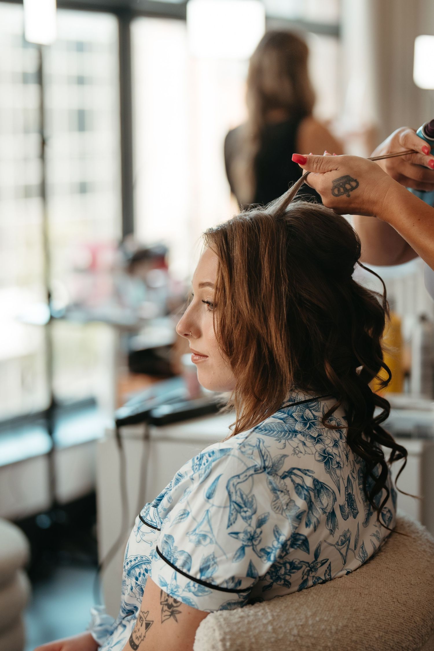 A hairstylist curling a young woman's long brunette hair in a bright salon, client wearing a blue floral robe.