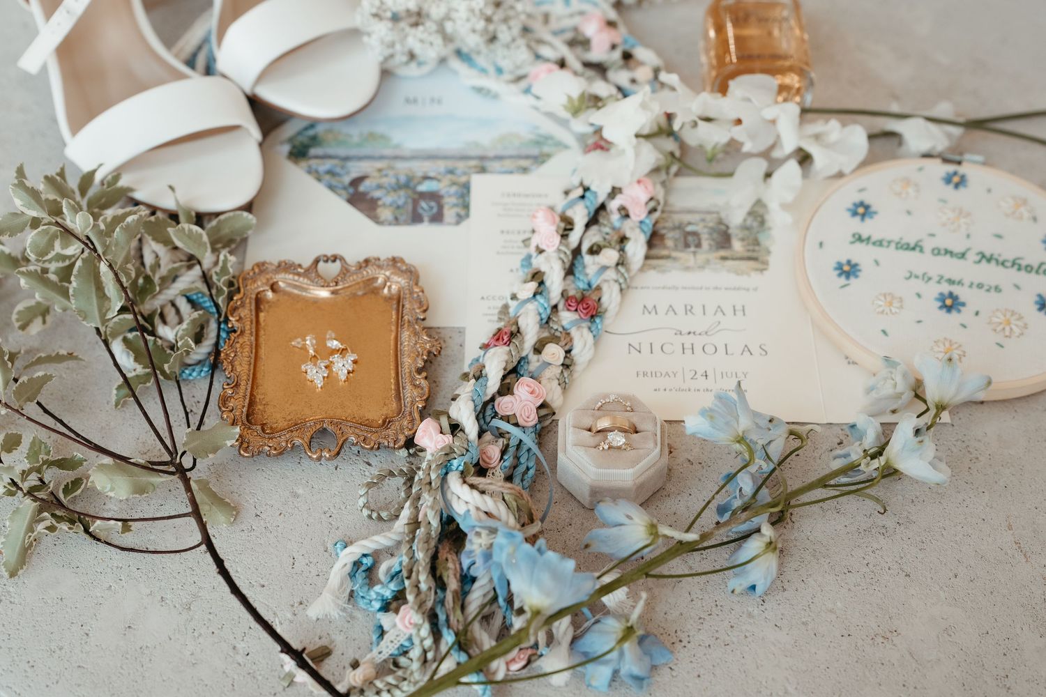 Wedding flat lay with white heels, floral invitation, ring box, braided cord, and delicate blue branches on neutral background.