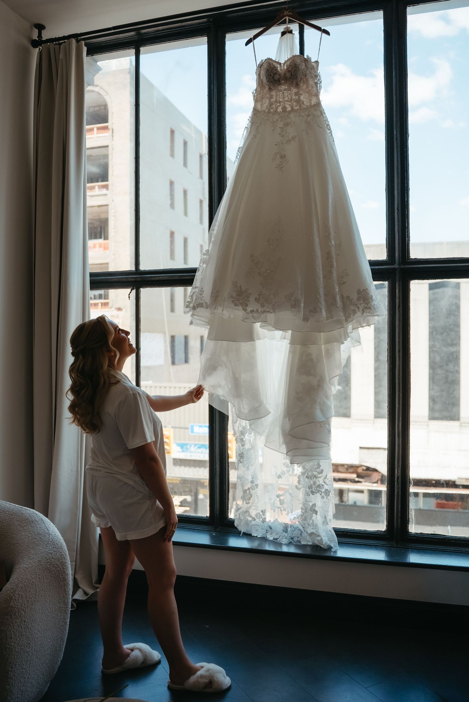 Bride in white robe admiring her lace ball gown wedding dress hanging by a large window in a hotel room.