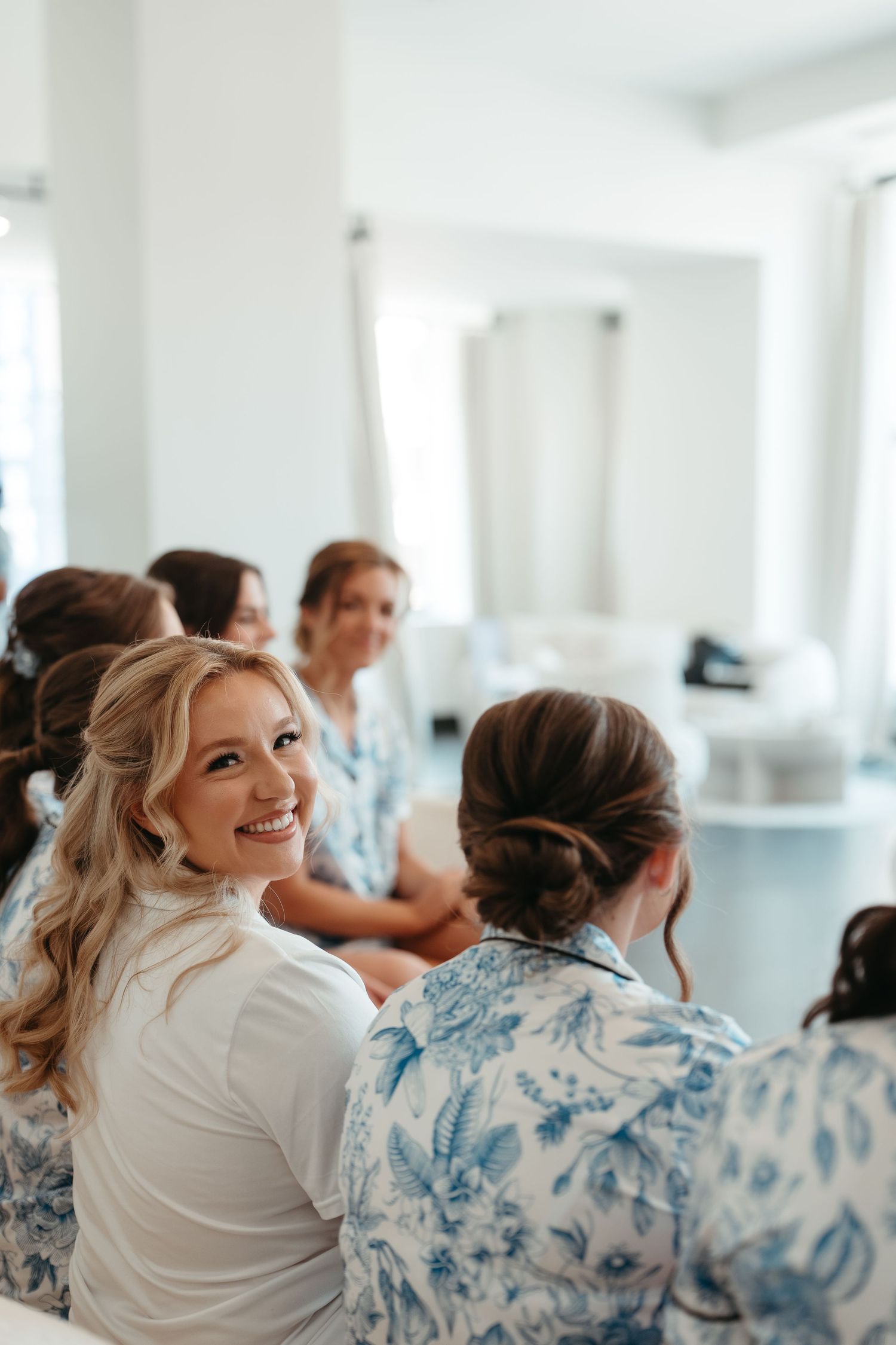 Smiling blonde bride in white robe with bridesmaids in blue floral robes getting ready for wedding day.