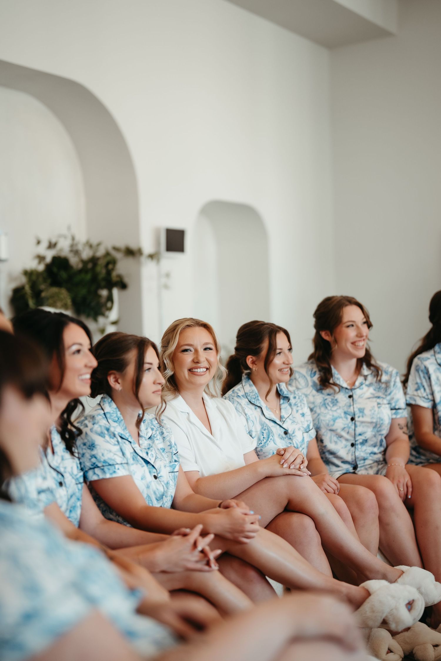 Bride and bridesmaids in matching blue floral pajamas sitting together smiling during wedding morning preparations.