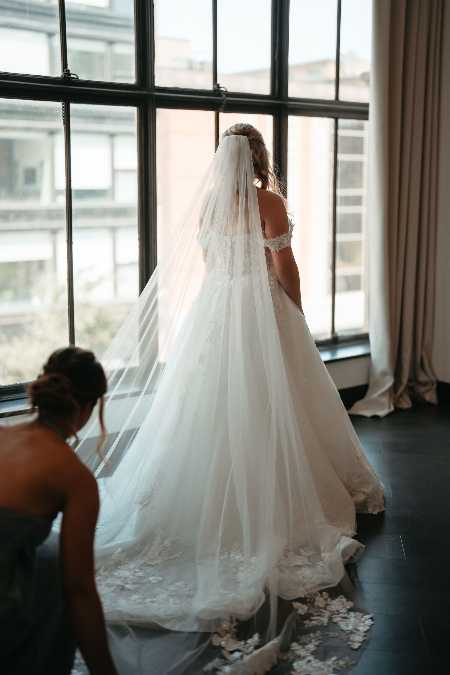 Bride in a stunning lace wedding gown with cathedral veil stands by tall windows as bridesmaid arranges her train.